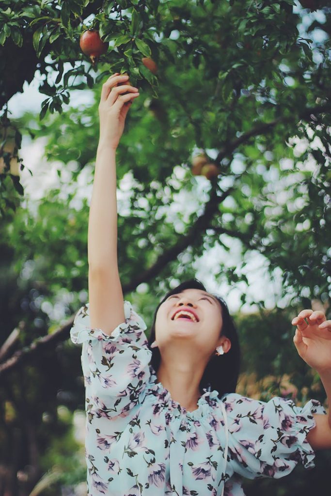 Smiling woman reaching for pomegranates on a sunny day, capturing joy and harvest.